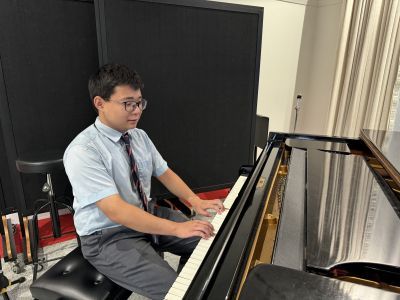 A young musician wearing a blue shirt and tie sits at a piano playing music.